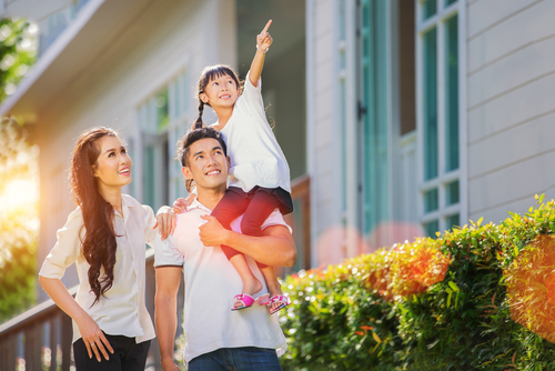 Dad holding daughter on shoulder in front of house while she points to sky. 